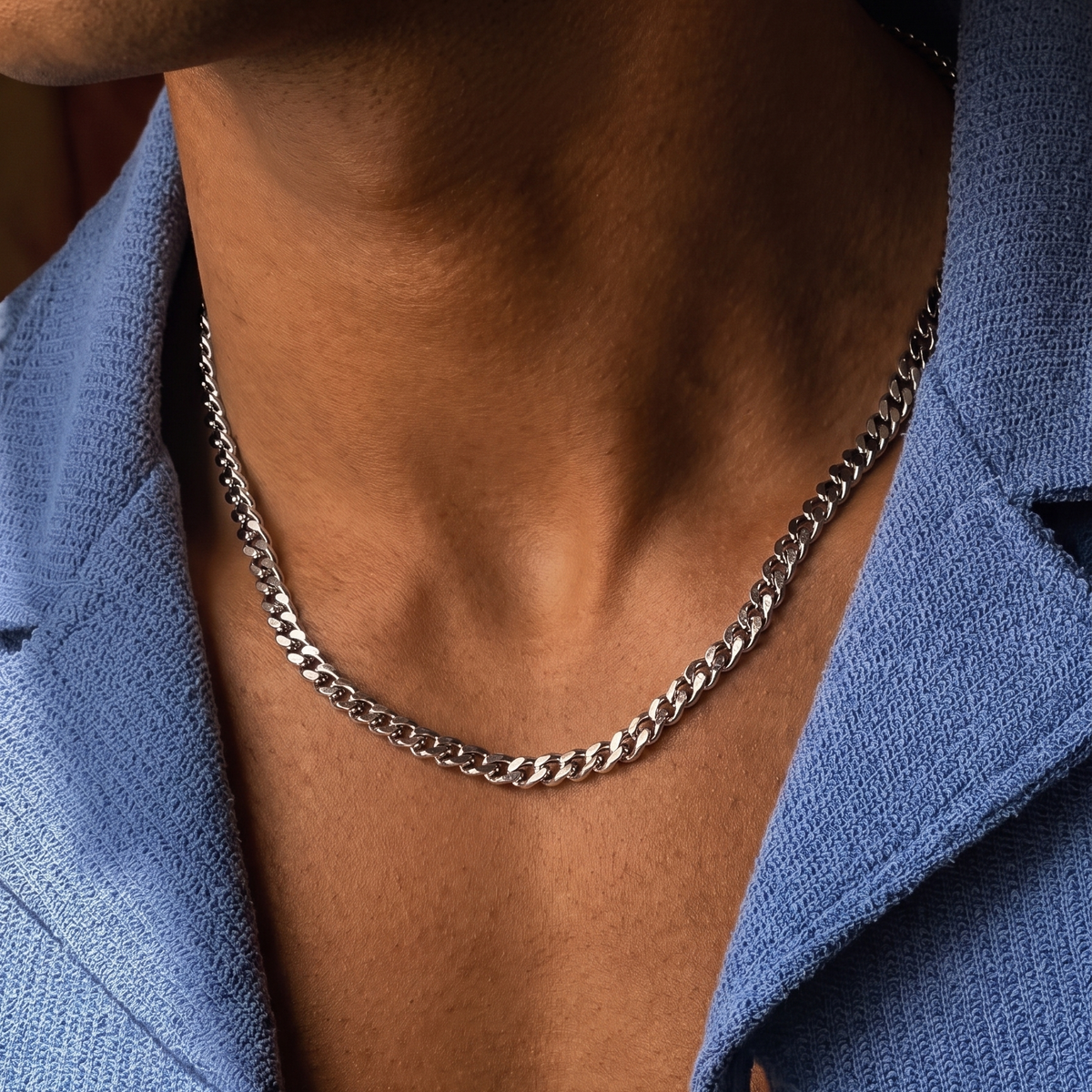 A close-up of a silver chain necklace worn by a person in a blue button-down shirt.