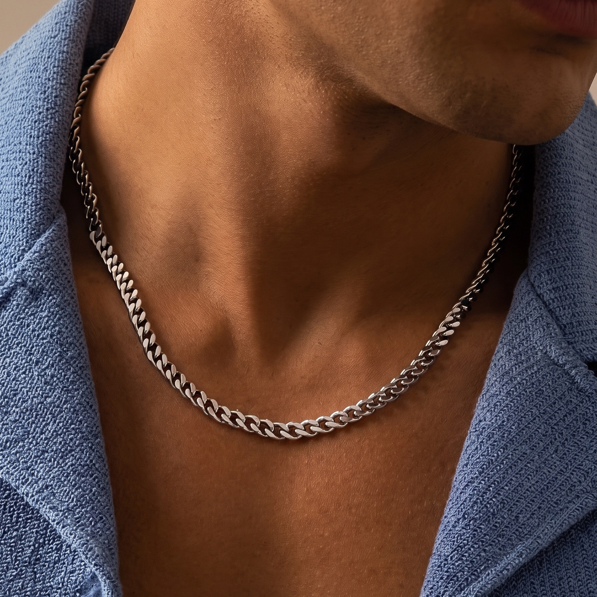 A close-up of a man's neck and chest wearing a silver-toned curb chain necklace over an open blue shirt.